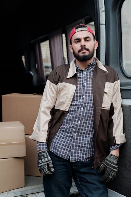 A young man with a beard, wearing a red baseball cap backwards, a checkered shirt, a beige and brown jacket, and grey work gloves, standing inside the back of a van during a home relocation. Behind hi