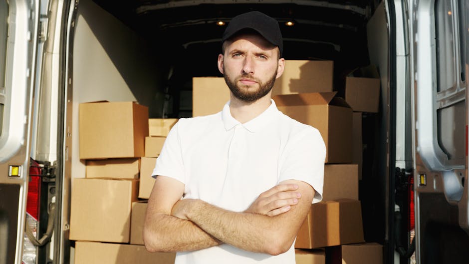 A young man with a beard, wearing a red baseball cap backwards, a checkered shirt, a beige and brown jacket, and grey work gloves, standing inside the back of a van during a home relocation. Behind hi