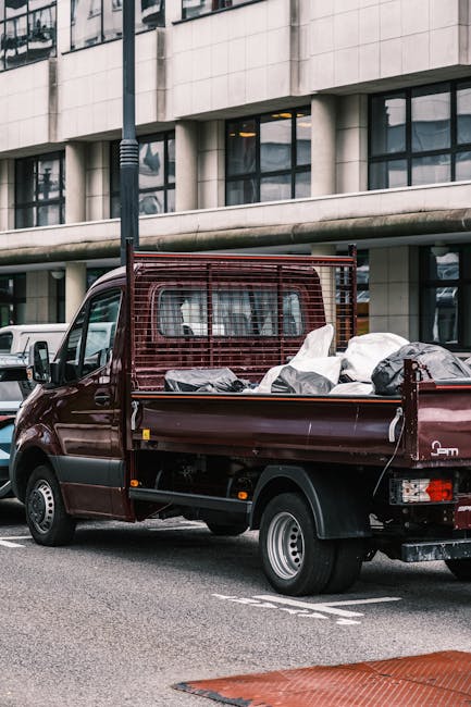 A burgundy flatbed van with a protective metal grille attached to the cab, parked on a city street outside a modern office building with large glass windows and concrete exterior. The van's cargo area