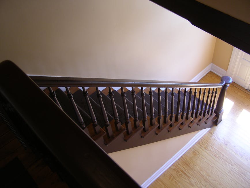 A top-down view of a spiral staircase inside a building, showing beige steps with dark non-slip strips along the edges, supported by a central metal pole and a brass-colored handrail. The staircase cu