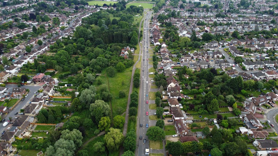 Aerial view of a residential neighbourhood along a busy main road, showing a line of cars and vans parked or moving in both directions. On one side of the road, there are terraced houses with paved front gardens, driveways, and small yards, while the other side features semi-detached houses with lush green front lawns and hedges. Green spaces and trees border the street, with some larger wooded areas visible further away. The scene captures the environment where house removals and furniture transport may take place, with the street clearly suitable for loading and unloading activities during a home relocation. Merton Man and Van operates in this area, providing professional removals and packing services for residents and businesses, as indicated by the page on SW19 moving routes and potential delays around Wimbledon Common to ensure smooth logistics during moving days.