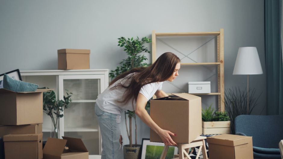 A man and a woman are inside a room filled with cardboard boxes used for house removals and packing, with some labeled with markers such as 'books'. The man, dressed in a grey hoodie, is sitting on th