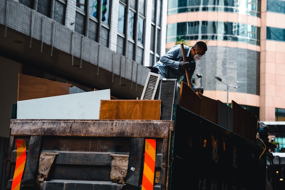 A man wearing a face mask and gloves is standing inside the back of a large, open-top skip truck, lifting a wooden cabinet with one hand while holding a tool in the other. The truck is parked on a paved street in an urban area, with modern high-rise buildings featuring glass and concrete facades visible in the background. Inside the skip, there are various household items and building materials, including cardboard boxes, plastic containers, a radiator, and pieces of wood. The loading process appears organized, with the man carefully handling the furniture during a home relocation or furniture transport. The scene reflects the typical logistics involved in removals services offered by Merton Man and Van, which include loading bulky waste and items during house removals or packing and moving to new premises.