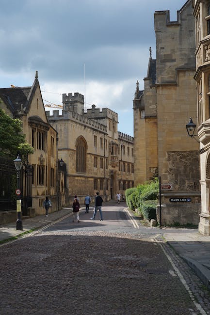 A narrow cobblestone street in a historic city centre with tall, ornate stone buildings on either side, including a large church or castle with battlements and arched windows visible in the background. Several pedestrians are walking along the street, some dressed in casual clothing, and the sky overhead is partly cloudy with patches of blue. The street features traditional black lampposts and greenery, including bushes lining the pavement. This setting illustrates an urban environment suitable for house removals and furniture transport, with the tight street space requiring careful loading and unloading of items, as might occur during a professional house move handled by Merton Man and Van.